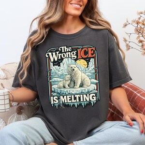 Puede incluir: Camiseta gris oscuro con un gráfico de dos osos polares en un témpano de hielo, con el texto "The Wrong Ice Is Melting". El diseño incluye un atardecer y un fondo de iceberg, creando un aspecto vintage.