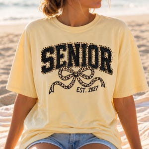 May include: A pale yellow short-sleeved t-shirt featuring the word SENIOR in black block letters with a leopard print fill. Below the text is a large leopard print bow graphic and the black text EST. 2027 printed on the front of the garment.