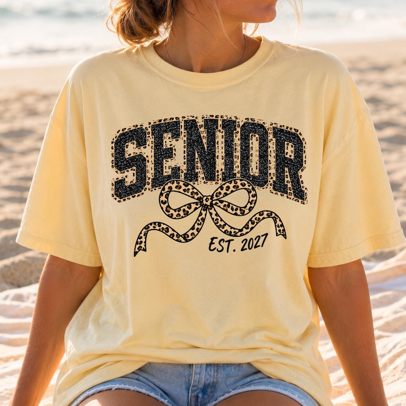 May include: A pale yellow short-sleeved t-shirt featuring the word SENIOR in black block letters with a leopard print fill. Below the text is a large leopard print bow graphic and the black text EST. 2027 printed on the front of the garment.