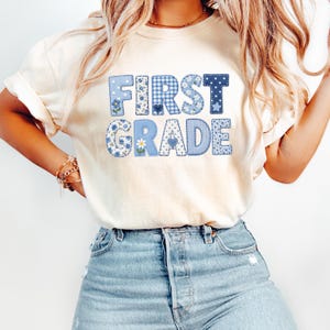 Pode incluir: Uma t-shirt de manga curta na cor creme com o texto FIRST GRADE em letras grandes de estilo bloco. Cada letra é preenchida com vários padrões azuis e brancos, incluindo xadrez vichy, bolinhas e estampas florais, com pequenos detalhes de corações.