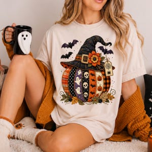Pode incluir: Uma t-shirt cor de creme com um gráfico de uma abóbora usando um chapéu de bruxa preto adornado com um girassol. A abóbora é decorada com padrões de fantasmas, caveiras e morcegos. Uma caneca preta com um design de fantasma é visível.