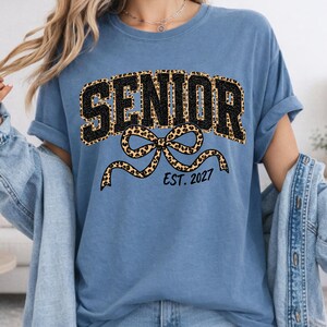 May include: A blue short-sleeved t-shirt featuring the word SENIOR in black block letters with a leopard print outline. Below the text is a large leopard print bow graphic and the black text EST. 2027 printed in a simple sans-serif font.
