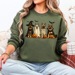 May include: An olive green crewneck sweatshirt featuring a graphic of four German Shepherd dogs dressed in Halloween costumes, including a witch, a ghost, a mummy, and a vampire, accompanied by small orange jack-o'-lanterns and black bats.