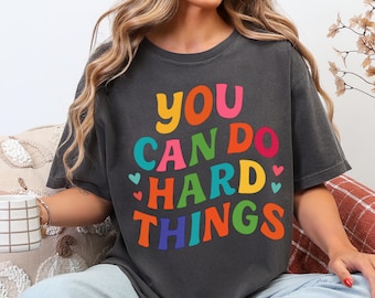 Może przedstawiać: Ciemnoszara koszulka z napisem "YOU CAN DO HARD THINGS" w kolorowych, bąbelkowych literach. Koszulka ma wygląd vintage, z efektem sprania i okrągłym dekoltem. Zawiera małe akcenty w kształcie serca.