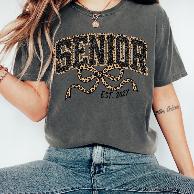 May include: A charcoal gray short-sleeve t-shirt featuring the word SENIOR in black block letters with a leopard print outline. Below the text is a large leopard print bow graphic and the black text EST. 2027 printed on the front of the shirt.