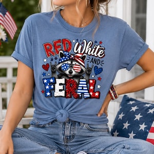Puede incluir: Camiseta azul claro con un dise&ntilde;o de mapache con gafas de sol y pa&ntilde;uelo rojo, blanco y azul. La camiseta tiene la frase "Red White and Feral" impresa, con estrellas y corazones.