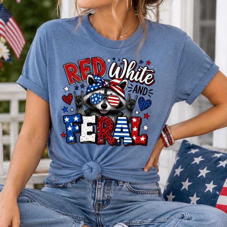Puede incluir: Camiseta azul claro con un dise&ntilde;o de mapache con gafas de sol y pa&ntilde;uelo rojo, blanco y azul. La camiseta tiene la frase "Red White and Feral" impresa, con estrellas y corazones.