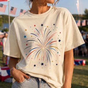 Peut inclure: T-shirt crème avec un motif de feu d'artifice coloré, orné de détails rouges, bleus et dorés, ainsi que d'étoiles. Ce haut décontracté à manches courtes est idéal pour les événements patriotiques.