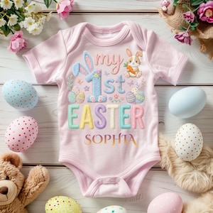 May include: A pale pink baby bodysuit featuring the text "my 1st EASTER SOPHIA" in pastel shades. The design includes a bunny, Easter eggs, and floral details. Decorated Easter eggs and a teddy bear surround the bodysuit.