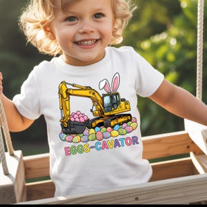 May include: White t-shirt with a yellow excavator design, complete with bunny ears, a bucket of pink eggs, and colourful Easter eggs. The text "EGGS-CAVATOR" is below. The shirt is worn by a child.