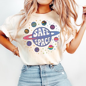 Puede incluir: Camiseta color crema con un gráfico colorido de un planeta con las palabras "SAFE SPACE" en una fuente retro. El planeta está rodeado de otros planetas y estrellas. La camiseta está hecha de un material suave.
