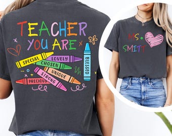 May include: Dark gray t-shirt with colorful text and crayon graphics. The back of the shirt reads "TEACHER YOU ARE" with words like "SPECIAL" and "LOVELY" on crayons. The front of the shirt has "MS. SMITT" and a pink heart.