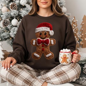 Może przedstawiać: Brązowa bluza z nadrukiem piernikowego ludzika w czapce Świętego Mikołaja i muszce. Piernikowy ludzik trzyma kubek z nadrukiem piernikowego ludzika i piankami. Część świątecznej kolekcji.