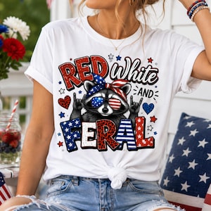 Puede incluir: Camiseta blanca con un dise&ntilde;o de un mapache con gafas de sol y una cinta patri&oacute;tica. El texto en la camiseta dice "Red, White, and Feral" en rojo, blanco y azul, con estrellas y corazones.