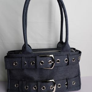 May include: A denim-colored handbag with two silver buckles and matching grommets. The bag has two handles and a zipper closure. The bag's design features a rectangular shape with a structured silhouette.