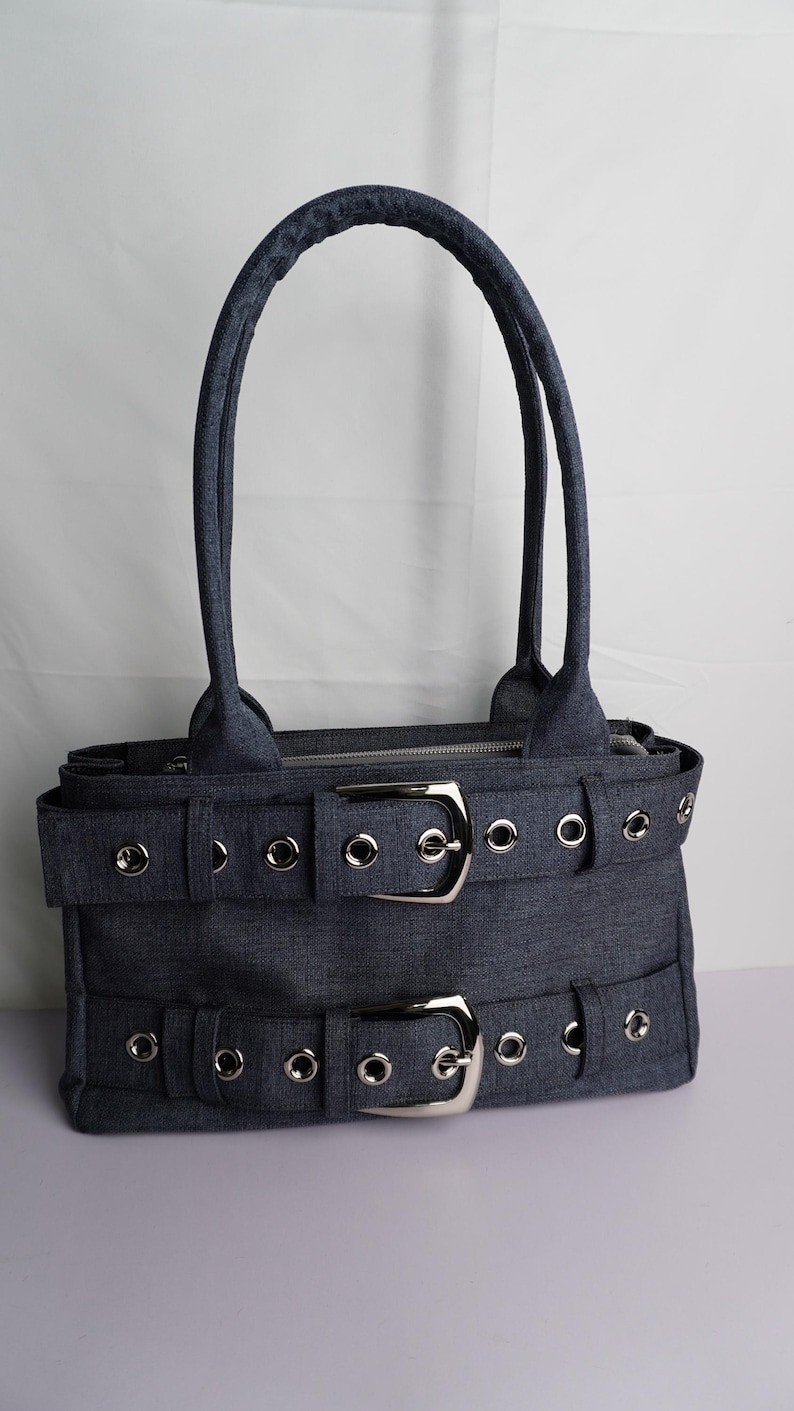 May include: A denim-colored handbag with two silver buckles and matching grommets. The bag has two handles and a zipper closure. The bag's design features a rectangular shape with a structured silhouette.