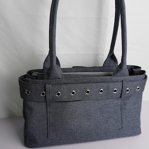 May include: A gray tote bag with two handles and a rectangular shape. The bag has a front pocket and is decorated with silver grommets. The bag is made of a textured fabric and is sitting on a white surface.