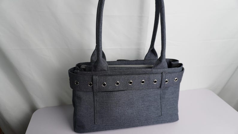 May include: A gray tote bag with two handles and a rectangular shape. The bag has a front pocket and is decorated with silver grommets. The bag is made of a textured fabric and is sitting on a white surface.
