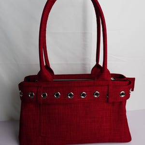 May include: A red tote bag with two handles and a rectangular shape. The bag features silver grommets along the top and a zippered closure. The material has a textured appearance.
