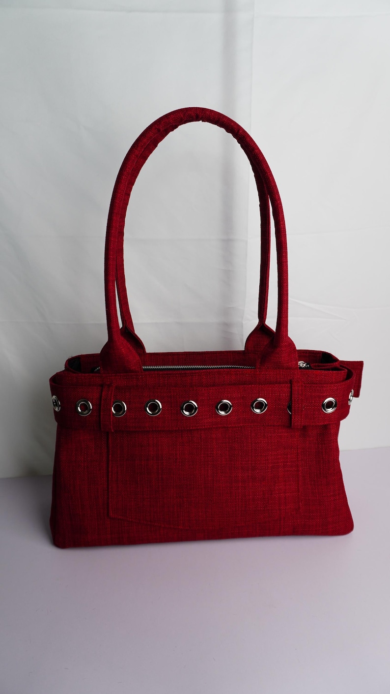 May include: A red tote bag with two handles and a rectangular shape. The bag features silver grommets along the top and a zippered closure. The material has a textured appearance.