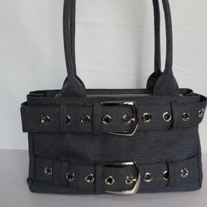May include: A gray tote bag with two handles. The bag features two horizontal straps with silver buckles and grommet details. The bag is made of a textured fabric and has a rectangular shape.