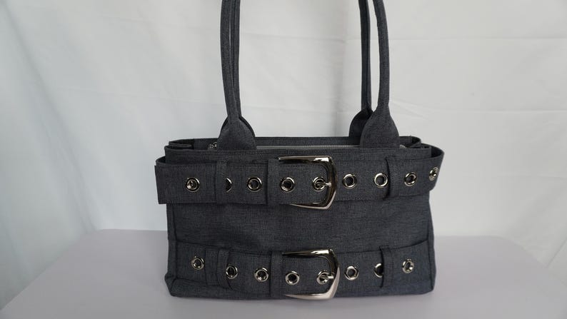 May include: A gray tote bag with two handles. The bag features two horizontal straps with silver buckles and grommet details. The bag is made of a textured fabric and has a rectangular shape.