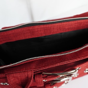 May include: A red handbag with a silver zipper and hardware. The bag has a rectangular shape with a top handle and a buckle closure. The interior is black.