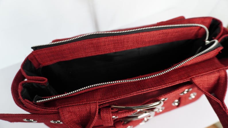 May include: A red handbag with a silver zipper and hardware. The bag has a rectangular shape with a top handle and a buckle closure. The interior is black.