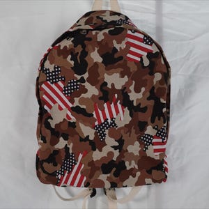 May include: A backpack featuring a camouflage pattern in shades of brown, tan, and black, interspersed with red and white stripes and white stars on a blue background. The bag has light-coloured straps and includes the text Youtube Tutorial and DDOL.