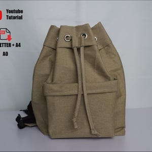 Può includere: Uno zaino beige con coulisse e una tasca frontale. La borsa ha occhielli metallici e una chiusura con coulisse. L'immagine include anche il testo "Youtube Tutorial" e "US_LETTER + A4 A0".
