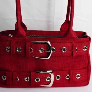 May include: A red handbag with two silver buckles and matching straps. The bag has a textured fabric and silver grommets. The bag has two handles and a zipper closure.