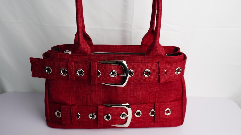 May include: A red handbag with two silver buckles and matching straps. The bag has a textured fabric and silver grommets. The bag has two handles and a zipper closure.