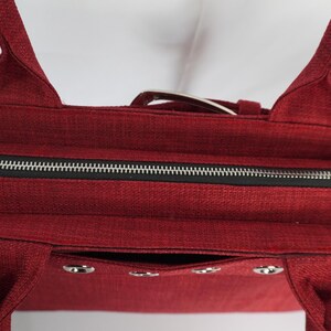 May include: A burgundy tote bag with a textured fabric. The bag features a black zipper, silver hardware, and two handles. The interior is black. The bag has a rectangular shape and a pocket on the front.