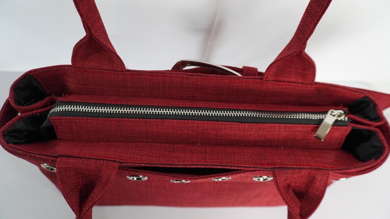 May include: A burgundy tote bag with a textured fabric. The bag features a black zipper, silver hardware, and two handles. The interior is black. The bag has a rectangular shape and a pocket on the front.