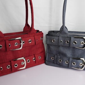 May include: Two tote bags, one red and one gray, with silver buckles and grommet details. Each bag has a rectangular shape, double handles, and a belt-like design with two large buckles. The bags are made of a textured fabric.