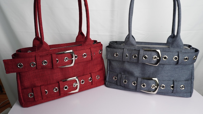 May include: Two tote bags, one red and one gray, with silver buckles and grommet details. Each bag has a rectangular shape, double handles, and a belt-like design with two large buckles. The bags are made of a textured fabric.