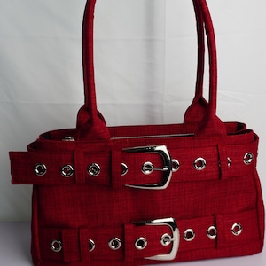 May include: A red handbag with two silver buckles and matching handles. The bag has a rectangular shape with a textured fabric and silver grommets. The bag is designed with two horizontal straps with silver buckles.