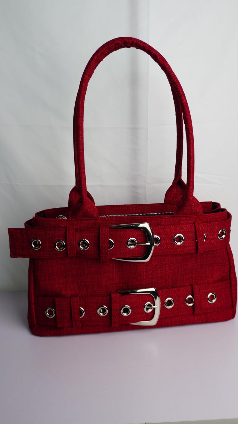 May include: A red handbag with two silver buckles and matching handles. The bag has a rectangular shape with a textured fabric and silver grommets. The bag is designed with two horizontal straps with silver buckles.