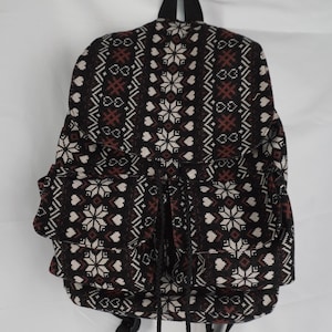 May include: A black backpack with a repeating pattern of white snowflakes, hearts, and geometric shapes in brown and white. The backpack has two side pockets and a drawstring closure.