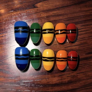 May include: A set of ten colorful, crayon-themed false nails. Each nail is painted in a different color, including blue, green, yellow, orange, and red, with black stripes. The nails are arranged on a wooden surface.