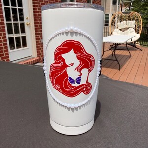 May include: A white cylindrical tumbler featuring a red graphic silhouette of a long-haired figure with a purple seashell top. The graphic is framed by a border of small white beads, with a vertical line of black beads on the left and right sides.