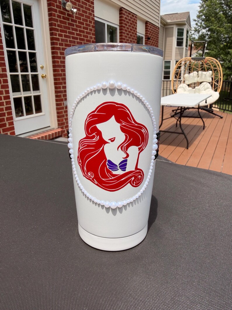 May include: A white cylindrical tumbler featuring a red graphic silhouette of a long-haired figure with a purple seashell top. The graphic is framed by a border of small white beads, with a vertical line of black beads on the left and right sides.