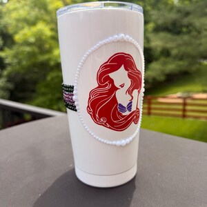 May include: A white stainless steel tumbler featuring a red-haired mermaid silhouette graphic enclosed in a white pearl border. The side of the tumbler is decorated with a horizontal band of small, multi-colored beads in black, purple, and pink.