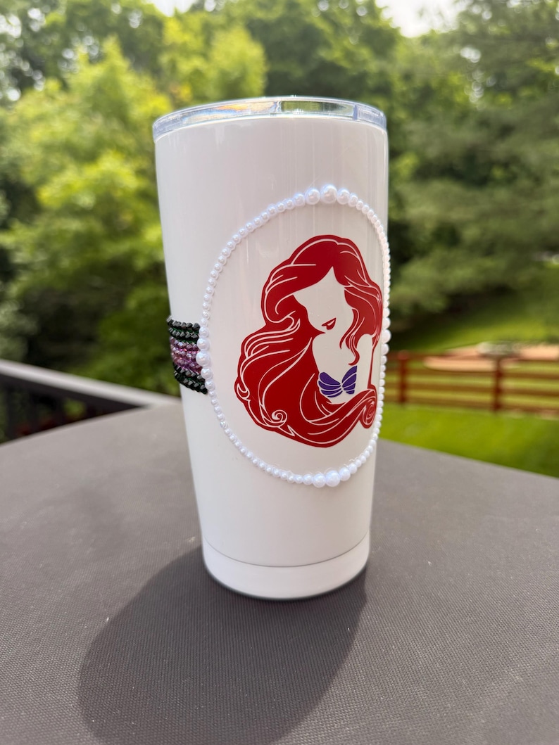May include: A white stainless steel tumbler featuring a red-haired mermaid silhouette graphic enclosed in a white pearl border. The side of the tumbler is decorated with a horizontal band of small, multi-colored beads in black, purple, and pink.