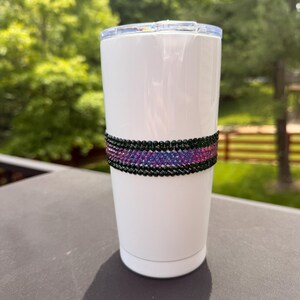 May include: A white cylindrical tumbler with a clear plastic lid, featuring a horizontal band around the center composed of rows of small, reflective black and iridescent purple rhinestones that create a textured, sparkling decorative accent.