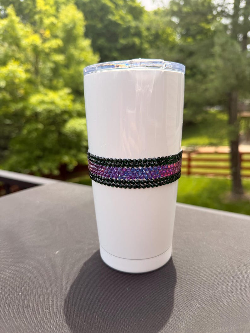 May include: A white cylindrical tumbler with a clear plastic lid, featuring a horizontal band around the center composed of rows of small, reflective black and iridescent purple rhinestones that create a textured, sparkling decorative accent.
