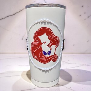 May include: A white stainless steel tumbler featuring a red silhouette graphic of a long-haired character with a purple seashell top. The graphic is framed by a circular border of small white faux pearls and accented by black beads on the sides.