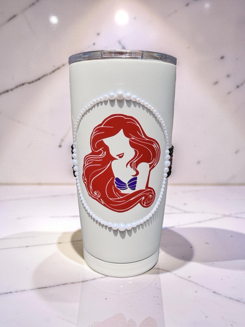 May include: A white stainless steel tumbler featuring a red silhouette graphic of a long-haired character with a purple seashell top. The graphic is framed by a circular border of small white faux pearls and accented by black beads on the sides.