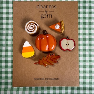 May include: A set of six autumn-themed mini magnets on a brown card labeled charms by gem and handmade mini magnets. The magnets include a cinnamon roll, a slice of pumpkin pie, a pumpkin, a candy corn, an apple slice, and an oak leaf.