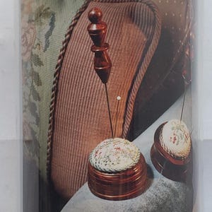 May include: A clear cylindrical plastic storage container holding a product with a dark blue label featuring the text For Creative Needlework and a white ribbon graphic. The label displays an image of two round pin cushions with wooden bases.